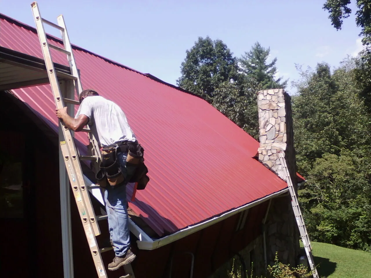 Roofer on a ladder beside a red metal roof and stone chimney — Soffit & Fascia Repair by Roofing Brothers in Starke